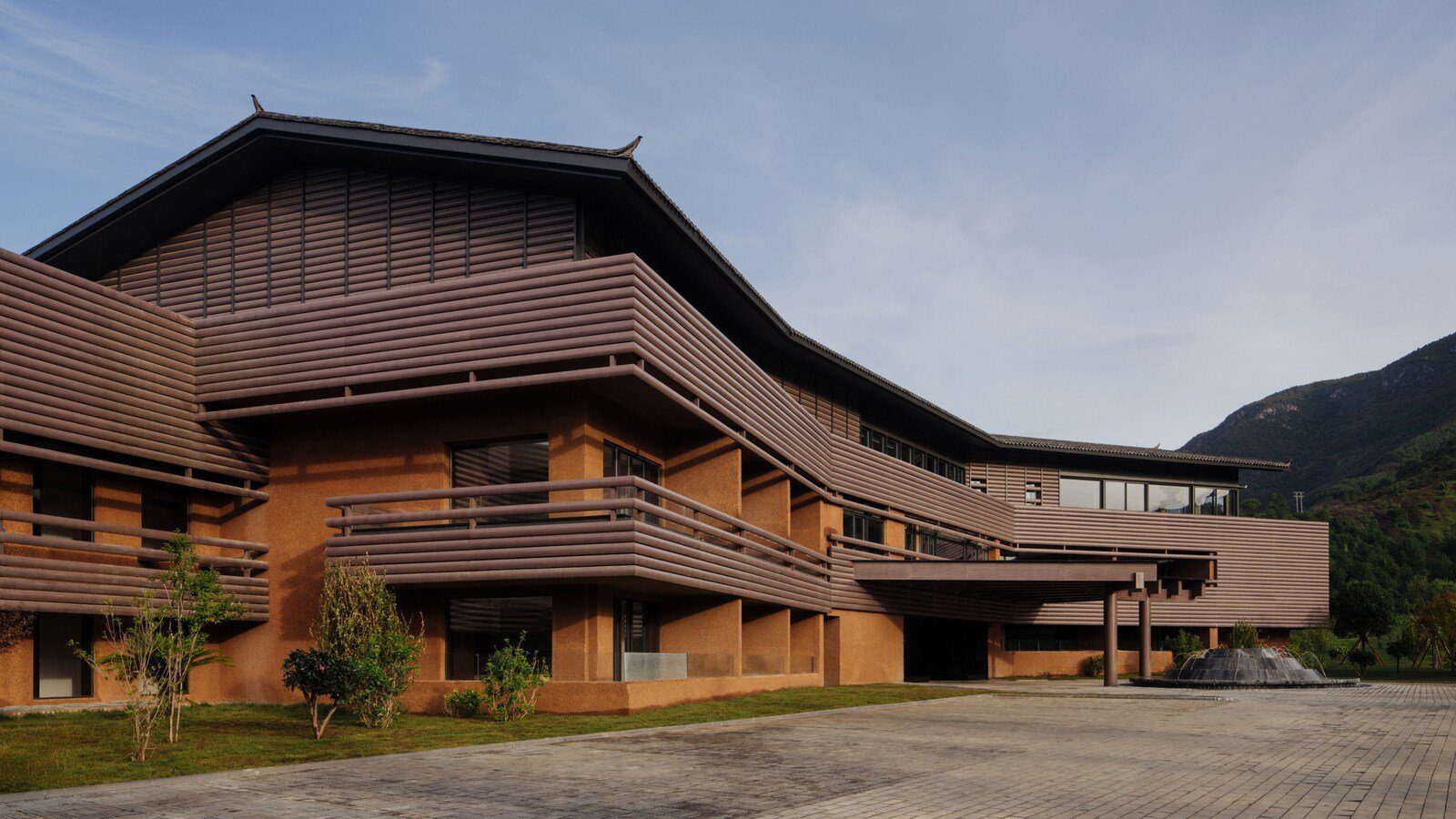 涼山文旅瀘沽湖隱奢逸境酒店的主樓與前方鋪石廣場,赭色牆面外是橫向的格柵與屋簷,右側有一座噴泉,背後是綠色山坡