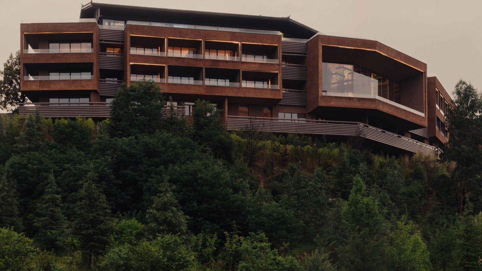 涼山文旅瀘沽湖隱奢逸境酒店的外觀,赭紅色的層疊建築立在林木茂密的山坡上,每層都有內凹的陽台,右側是整面玻璃的大空間
