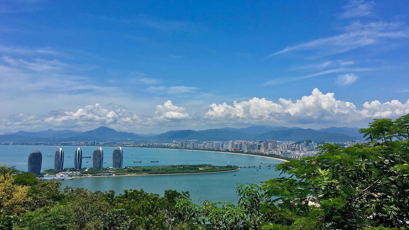 周邊景色:從鹿回頭一帶的山上俯瞰三亞灣,海上人工島立著五棟高樓,沿岸是城市樓群,後方是連綿的山