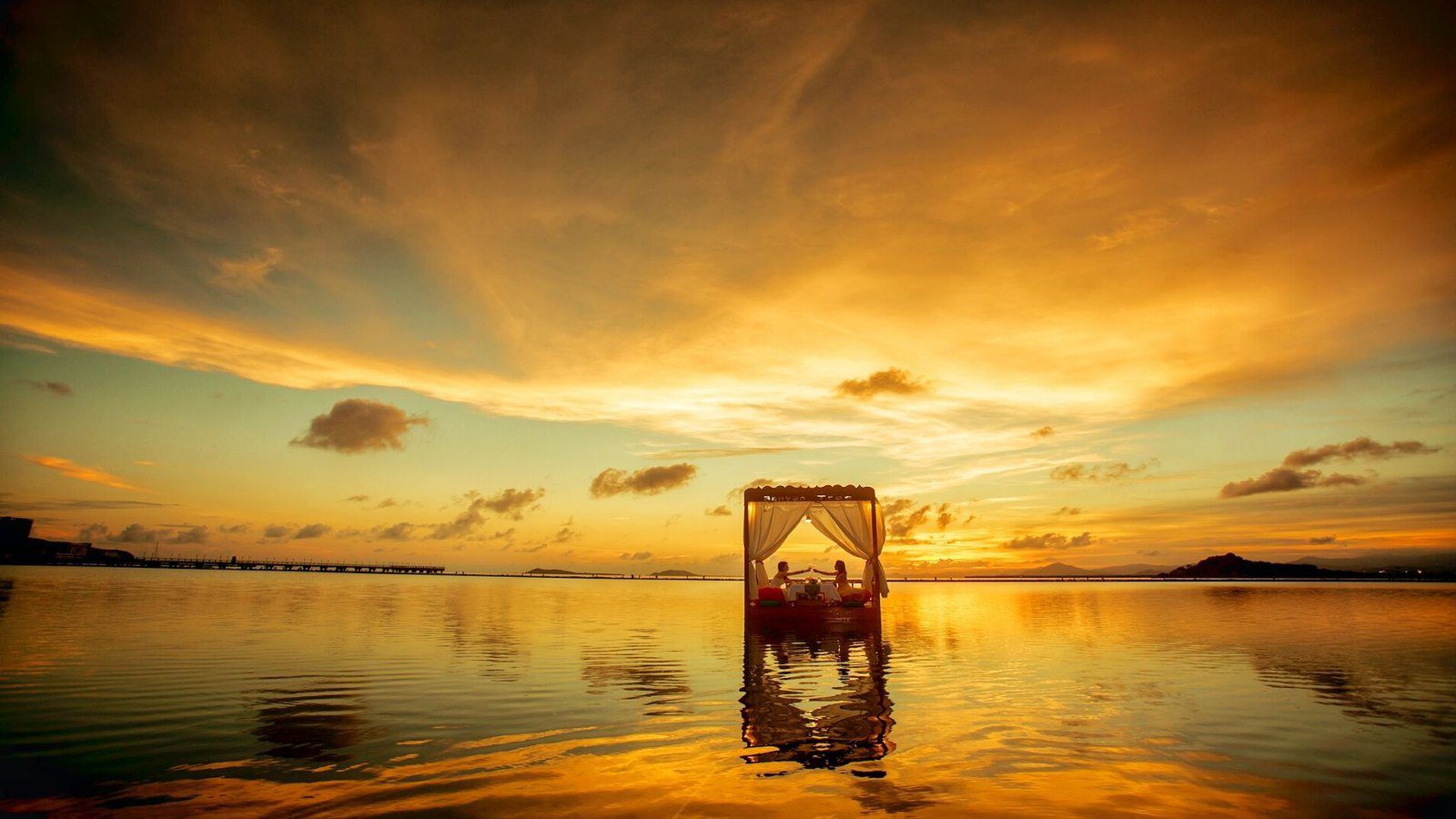 三亞悅榕莊的海上漂浮晚餐，金黃色夕陽下一座掛著白紗的四柱平台浮在平靜海面上，兩人在平台裡用餐