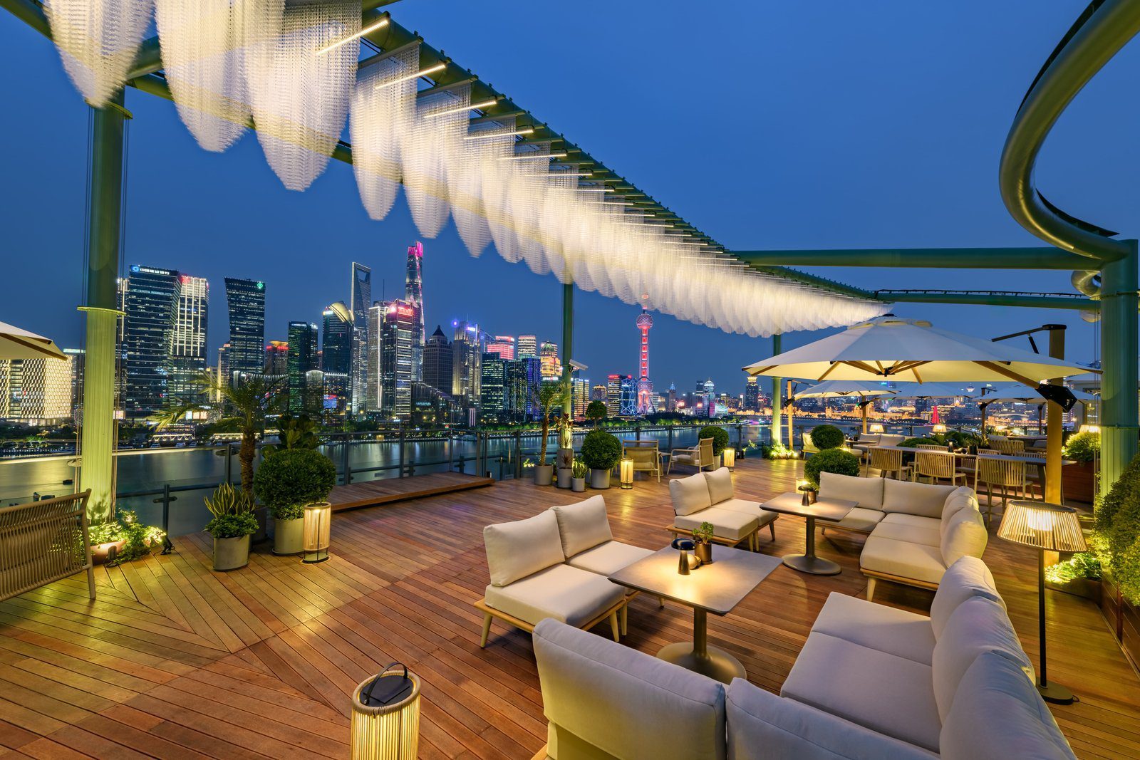 上海外灘悅榕莊 TALES SHANGHAI 屋頂露台的夜景，木地板上是白色沙發與遮陽傘，上方廊架垂著白色燈飾，欄杆外是黃浦江與陸家嘴