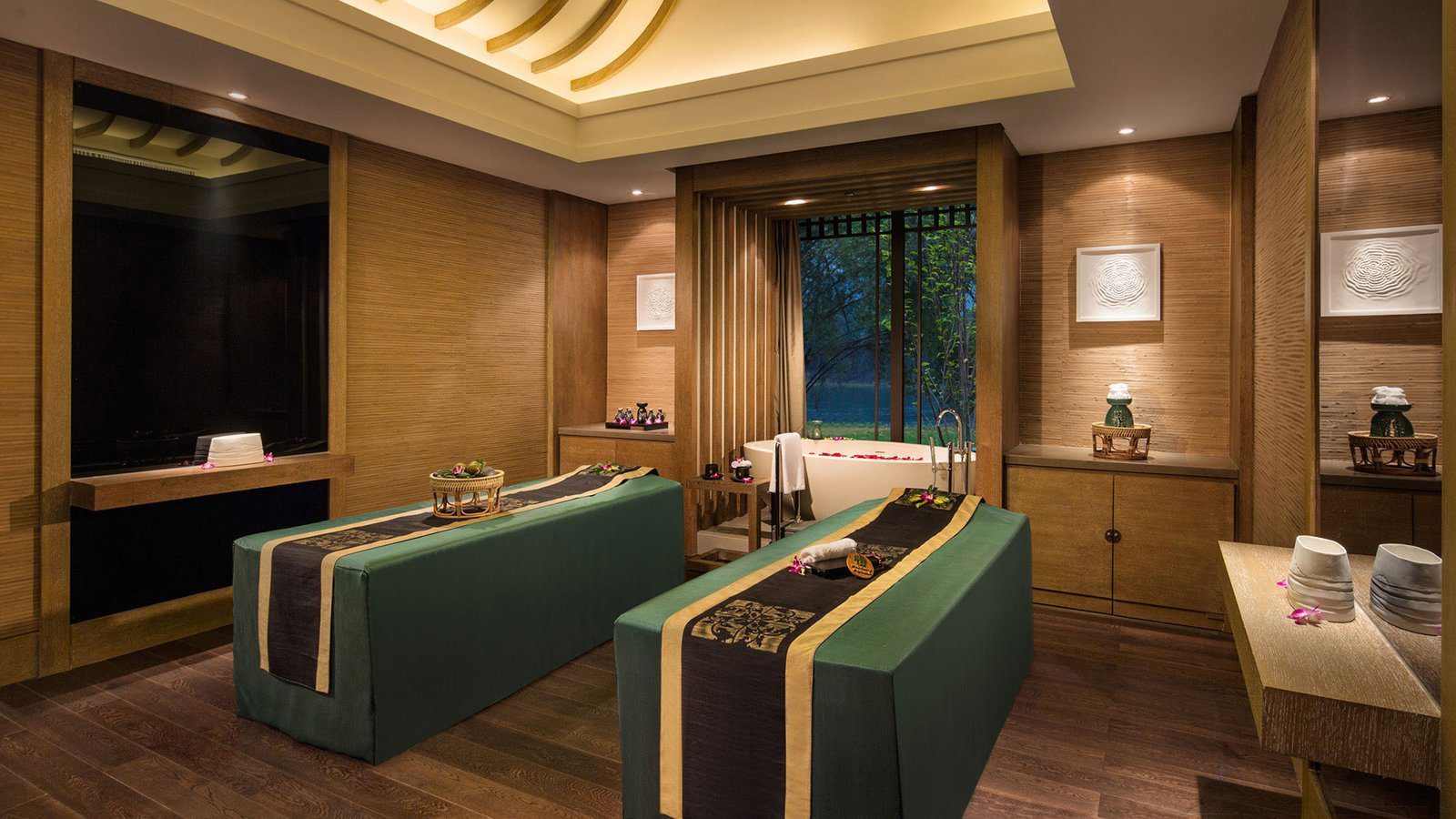 陽朔悅榕莊悅榕 SPA 的雙人療程室，兩張墨綠色按摩床並排，後方窗前擺著一座白色浴缸，牆面是木紋與編織壁材