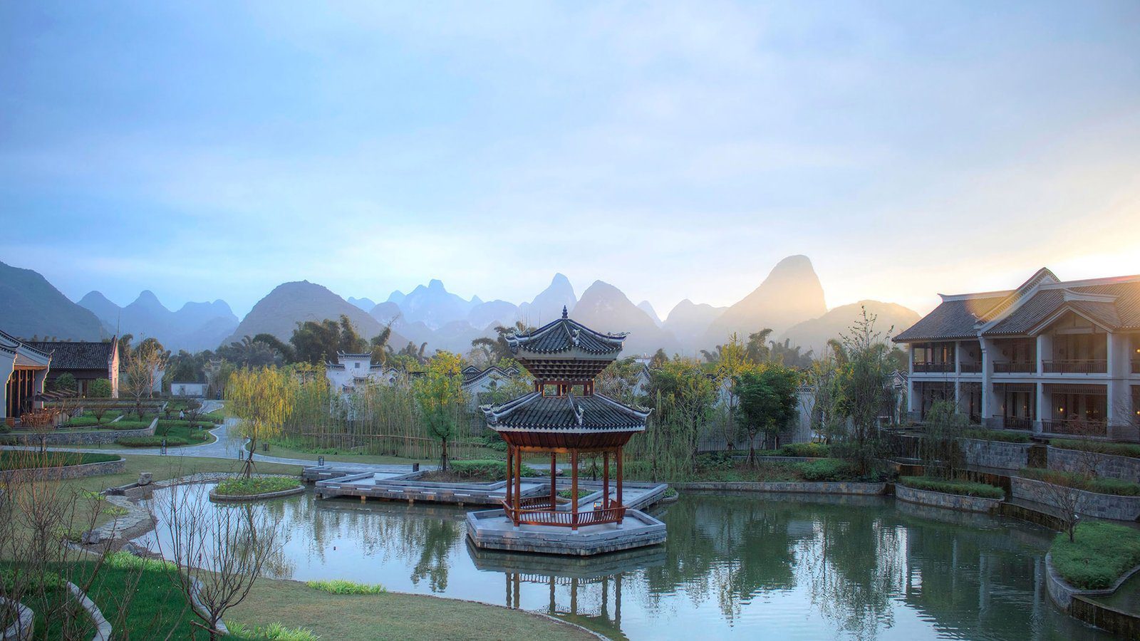 陽朔悅榕莊園區裡的湖面，湖心立著一座紅柱雙層涼亭，右側是兩層樓的客房建築，遠方是一整排喀斯特山峰