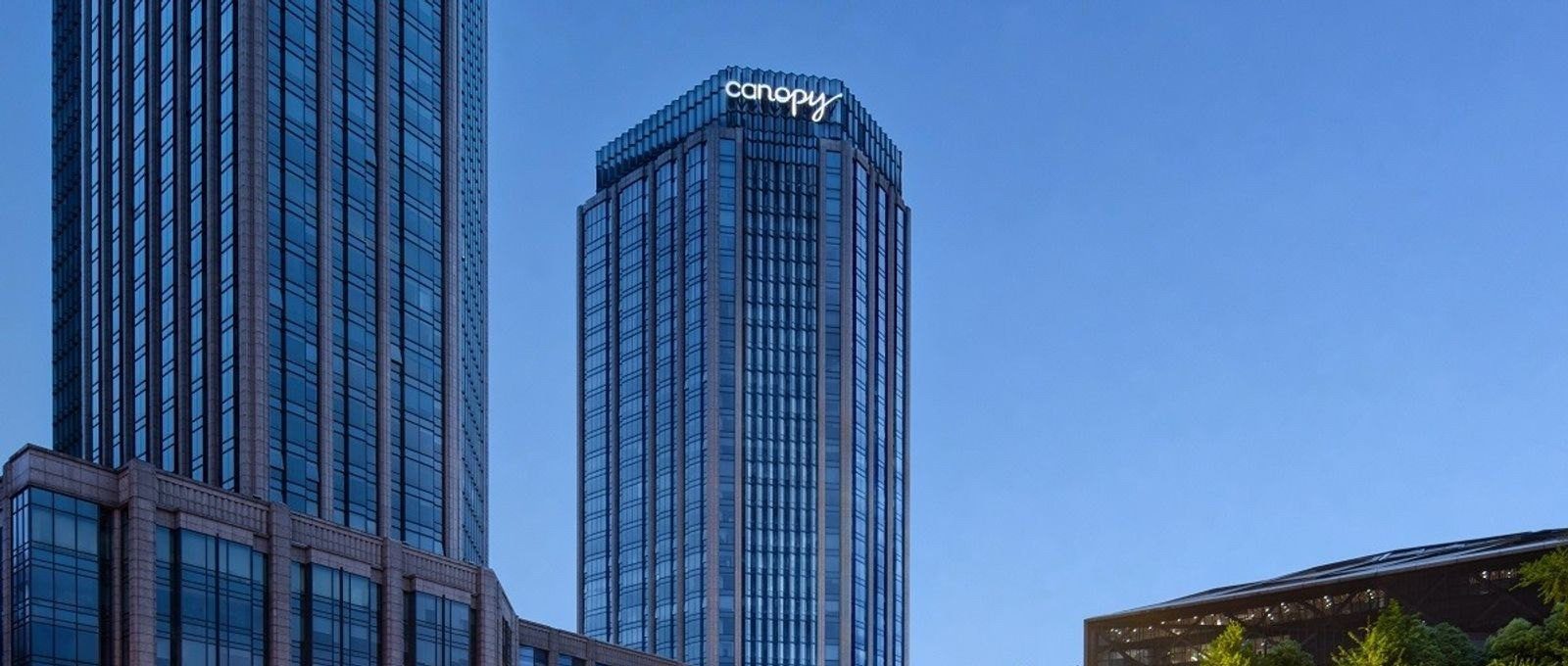 成都領地希爾頓嘉悅里酒店所在的塔樓在藍色天空下，樓頂亮著白色 canopy 招牌，左側是另一棟玻璃帷幕高樓