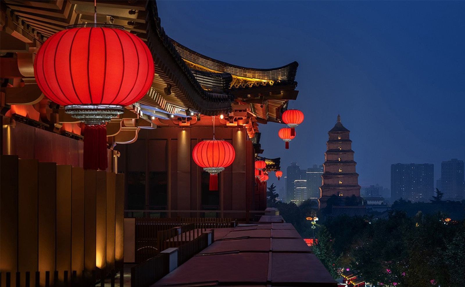 夜間的唐風屋簷下垂掛紅燈籠,遠方是亮燈的大雁塔與城市高樓(周邊景觀)
