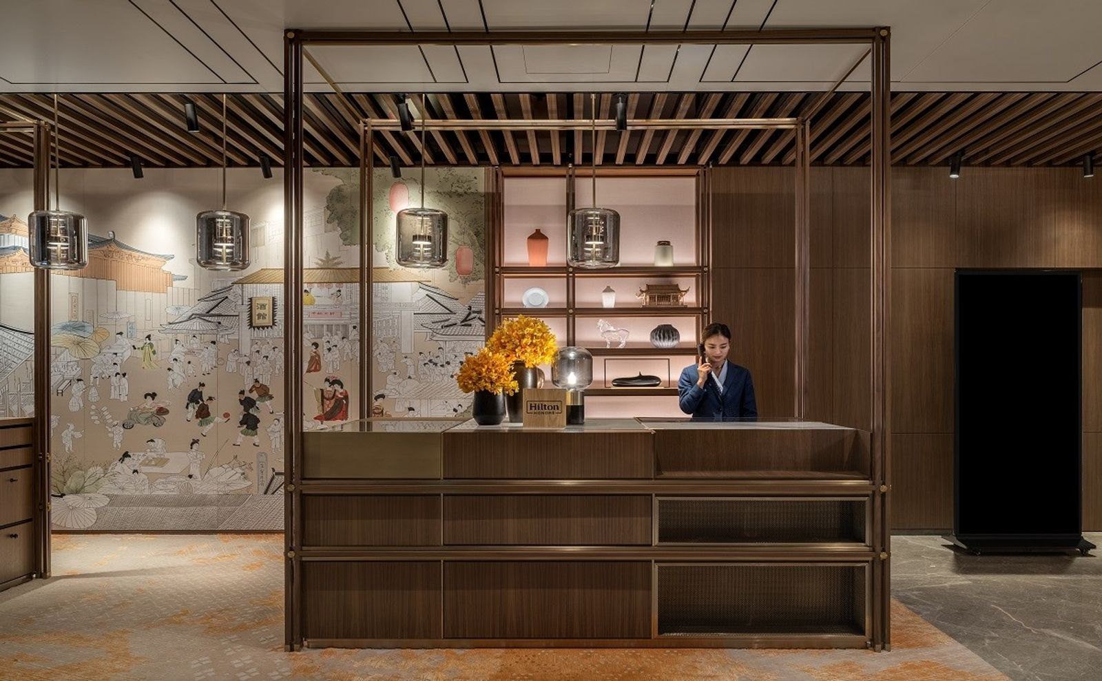 西安曲江希爾頓嘉悅里酒店的接待台,金屬框架下是木質櫃檯與黃色花束,左側牆面是唐代市集風格壁畫