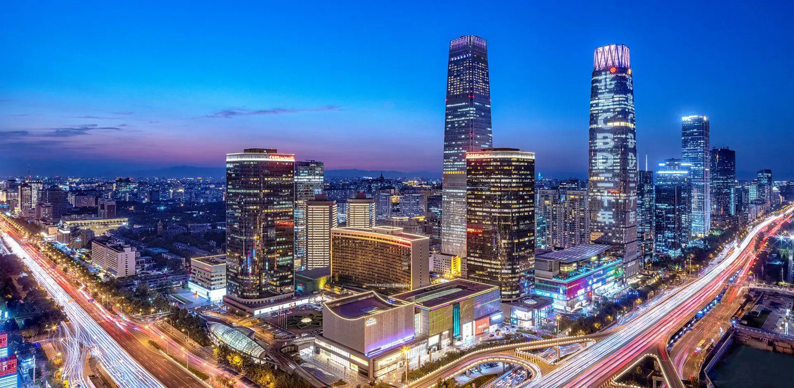 國貿一帶的夜景空拍，高樓群之間是亮燈的商場建築，兩側高架道路上拉出車流光軌