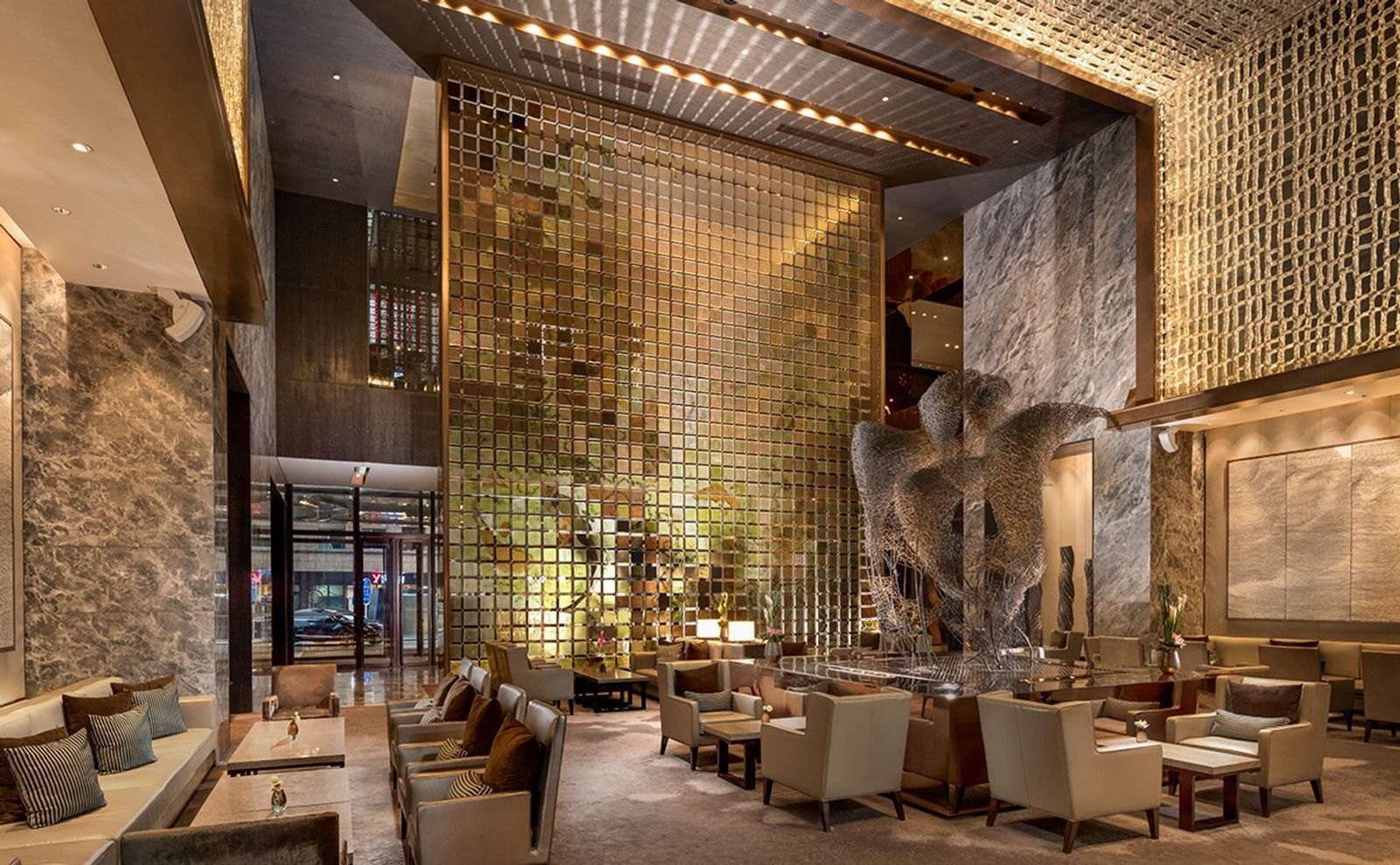 北京康萊德酒店挑高的大堂酒廊，中央立著金色方格屏風與大型金屬雕塑，四周擺著米色扶手椅與矮桌