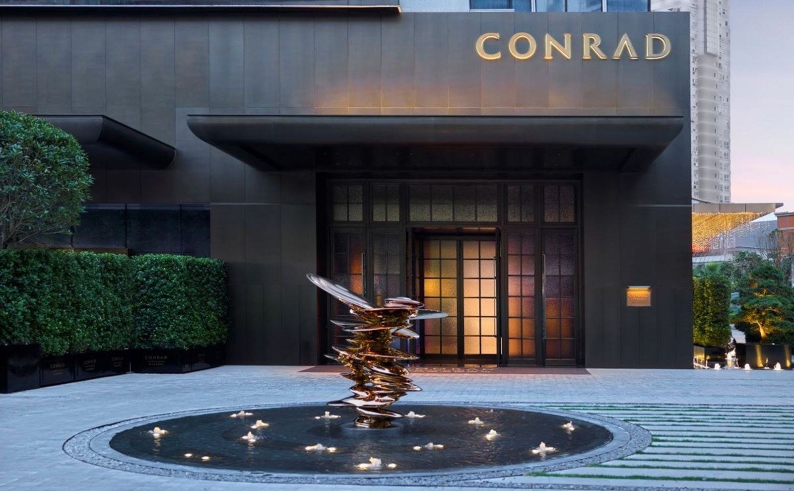 重慶康萊德酒店地面入口，深色外牆上是金色 CONRAD 字樣，門前水池中立著一座金屬雕塑