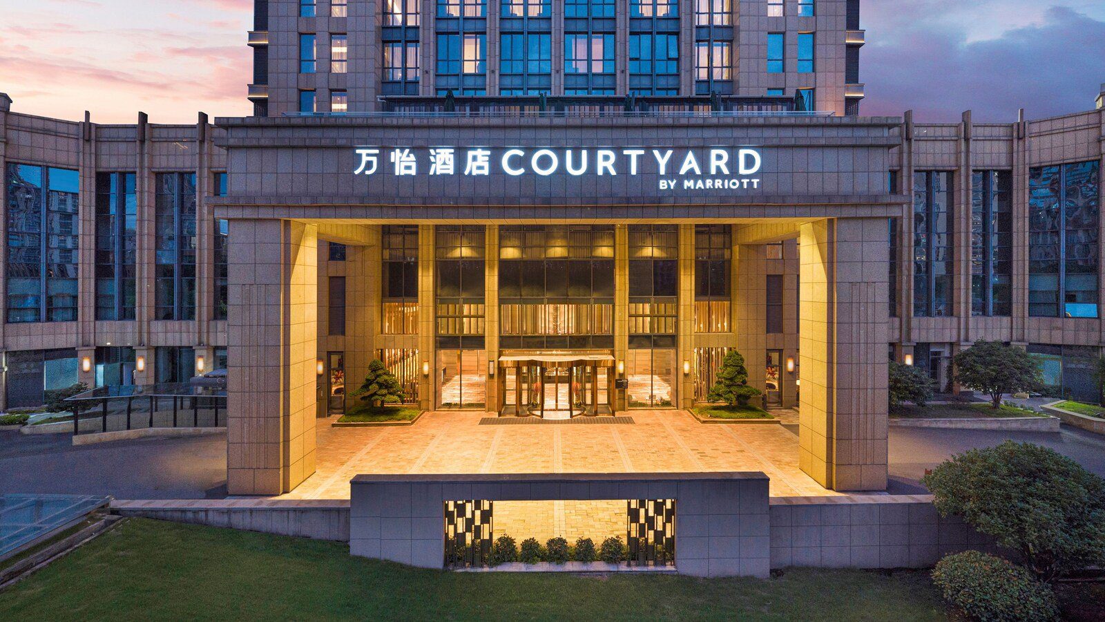 杭州蕭山萬怡酒店黃昏時的入口門廊，上方亮著 COURTYARD BY MARRIOTT 招牌，中央是旋轉門，前方是草坪
