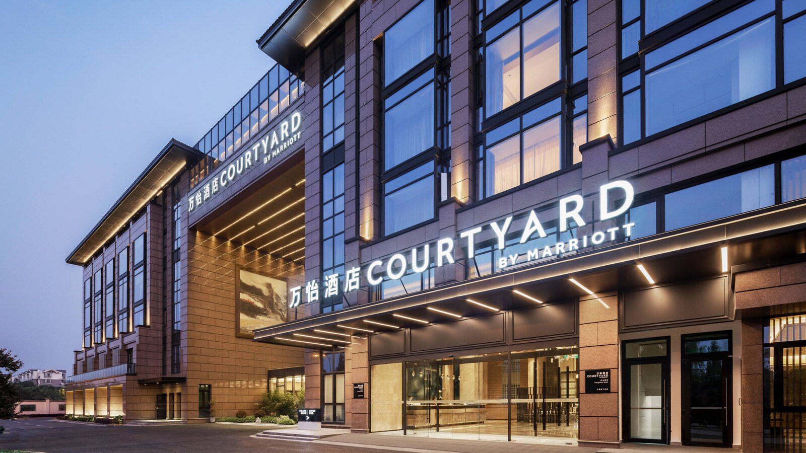 杭州西溪萬怡酒店黃昏時的入口,雨遮上方亮著 COURTYARD BY MARRIOTT 招牌,玻璃門內是大堂