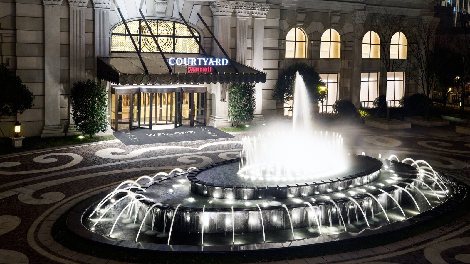 上海蘇寧環球萬怡酒店的入口夜景，雨遮上是 COURTYARD Marriott 招牌，門前有亮燈的圓形噴泉