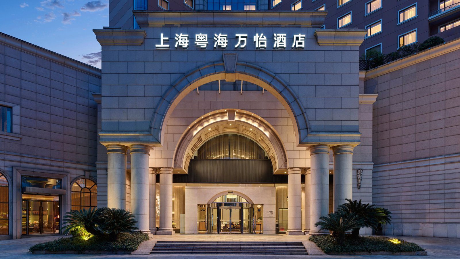 上海粵海萬怡酒店主入口,石砌拱門與成排圓柱,上方是亮燈的館名招牌,門前台階兩側種著蘇鐵