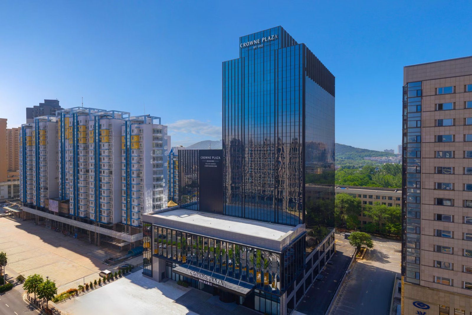 大連凱倫皇冠假日酒店空拍外觀，玻璃塔樓頂端有 CROWNE PLAZA 字樣，後方是綠色山丘
