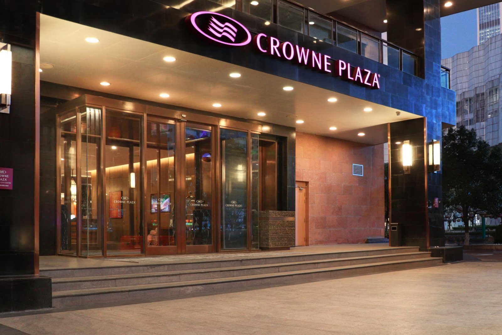 昆明中心皇冠假日酒店入口雨遮,上方亮著 CROWNE PLAZA 招牌,台階後是玻璃旋轉門與大堂燈光