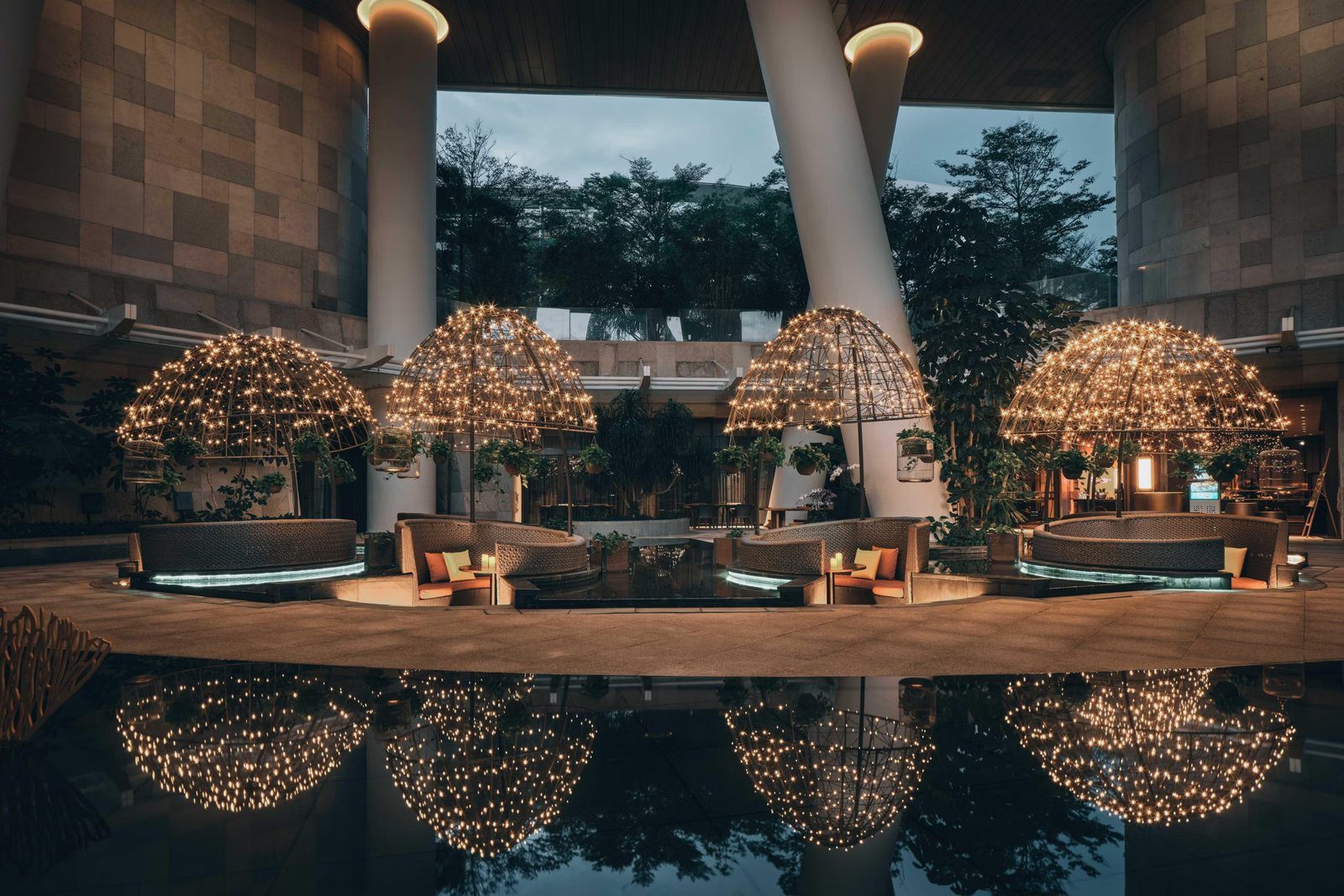 夜晚大堂挑空處的水池邊，四座掛滿燈串的圓頂造型下擺著弧形沙發，倒影映在水面上