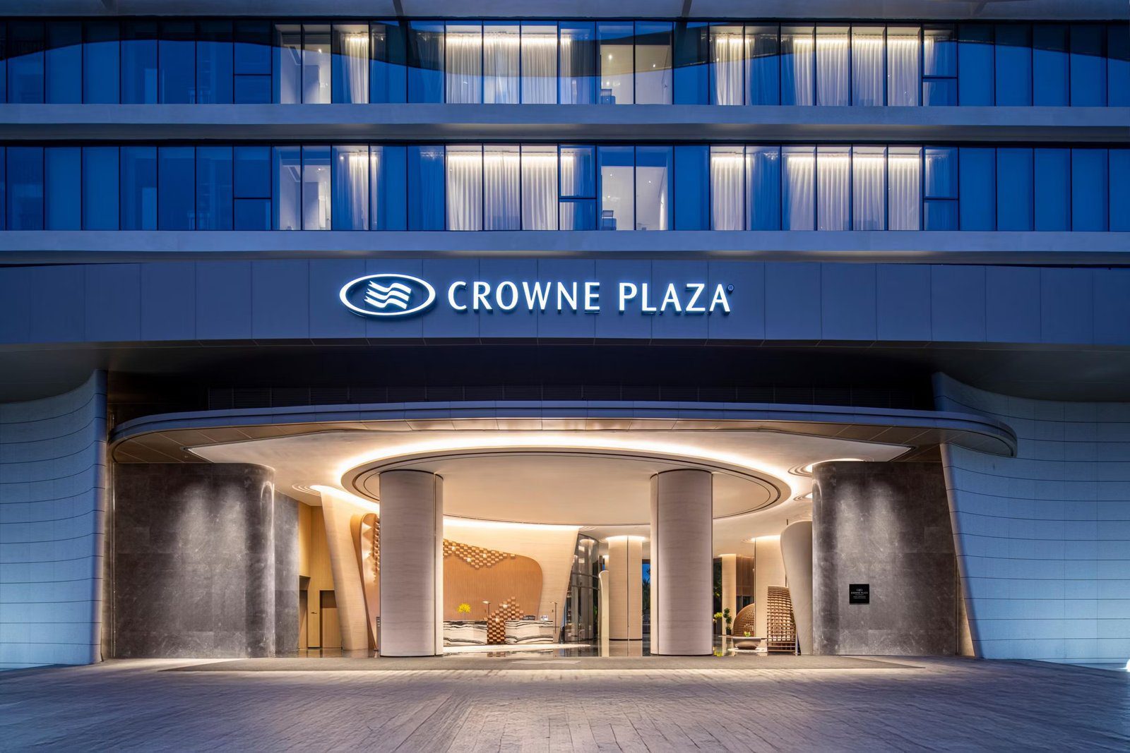 夜間大門上方亮著 CROWNE PLAZA 招牌，弧形雨遮下的兩根圓柱後方是大堂