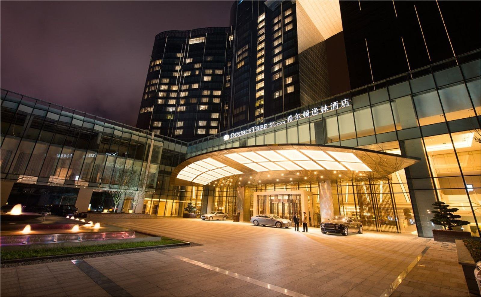 夜間的安順百靈希爾頓逸林酒店正門，亮燈的弧形雨庇上方是 DoubleTree by Hilton 與中文館名招牌，門前停著轎車，左側有噴泉水池