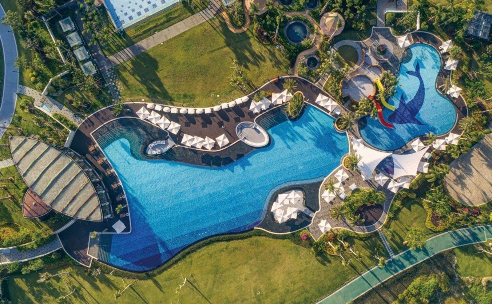 正上方空拍蜿蜒的大型泳池,池邊排滿白色遮陽傘,右側是底部鋪著海豚圖案、有彩色滑梯的戲水池