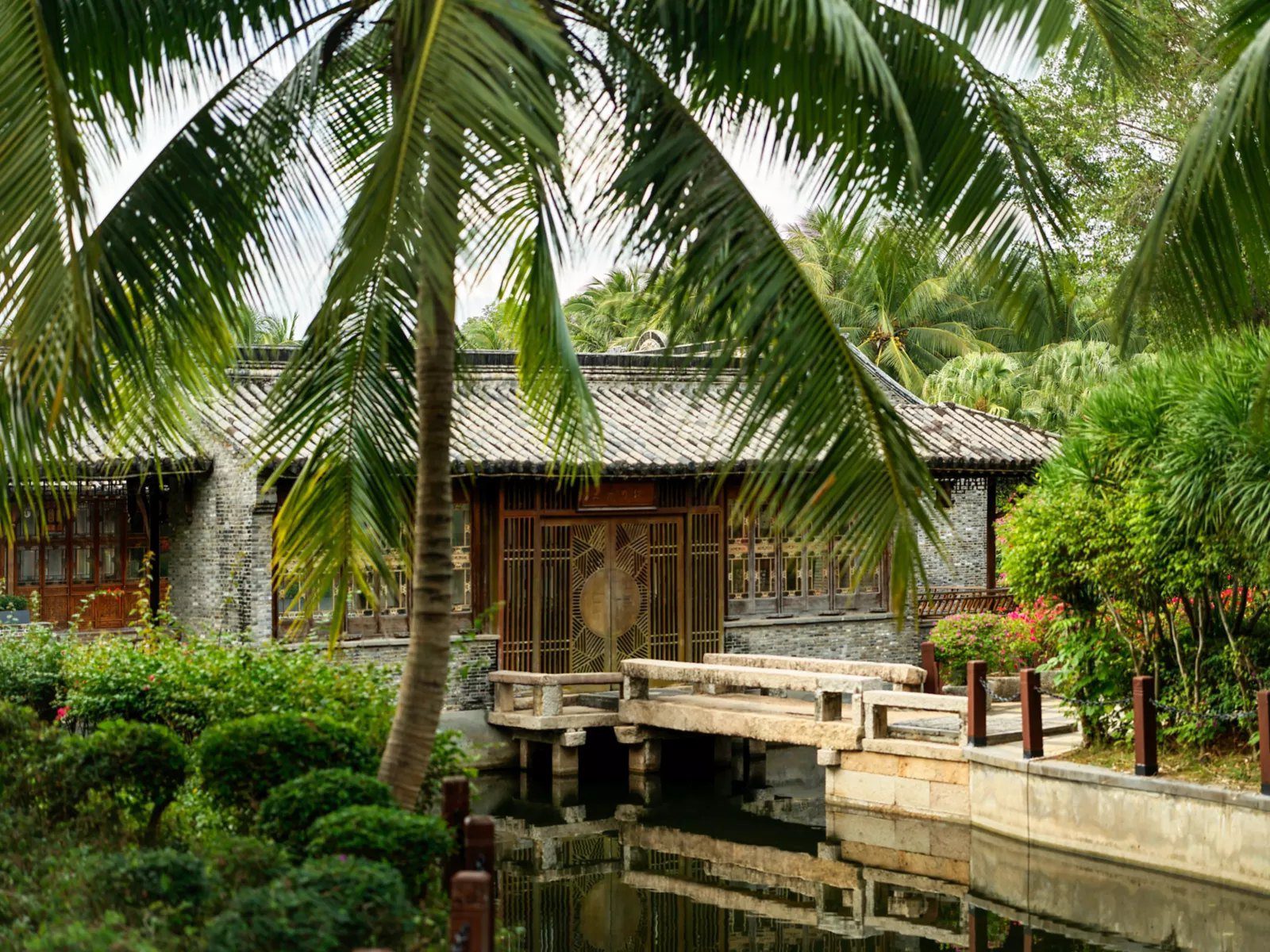 三亞海棠灣開維費爾蒙酒店園區裡的青磚灰瓦老建築，木格門窗前有一座石板橋跨過水道，前景是椰子樹與灌木