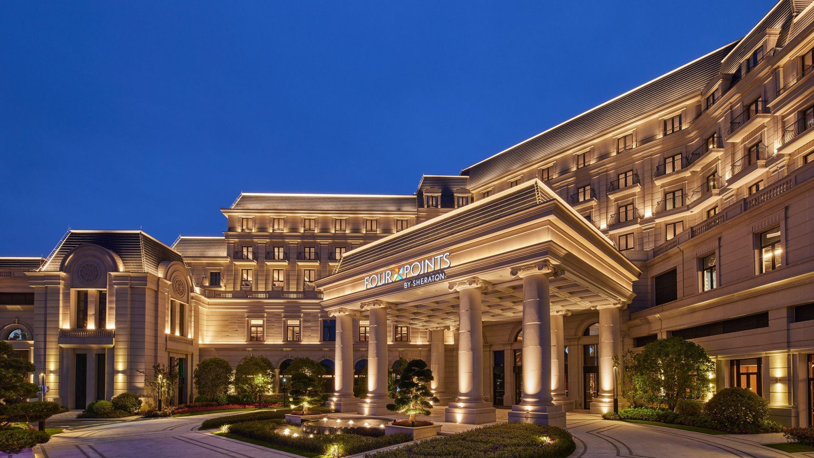 上海楓涇福朋喜來登酒店的入口門廊夜景,羅馬柱門廊上方是 FOUR POINTS BY SHERATON 招牌,前方是造景松樹與水景