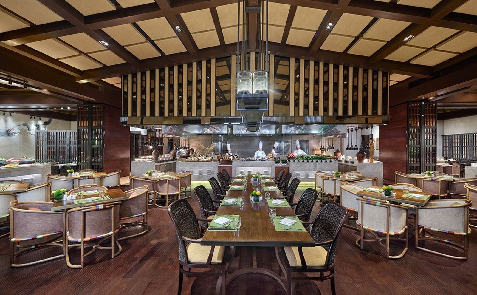 大理實力希爾頓酒店的餐廳用餐區，木格天花板下擺著長餐桌與圓背扶手椅，後方是開放式廚房，廚師在檯前備餐