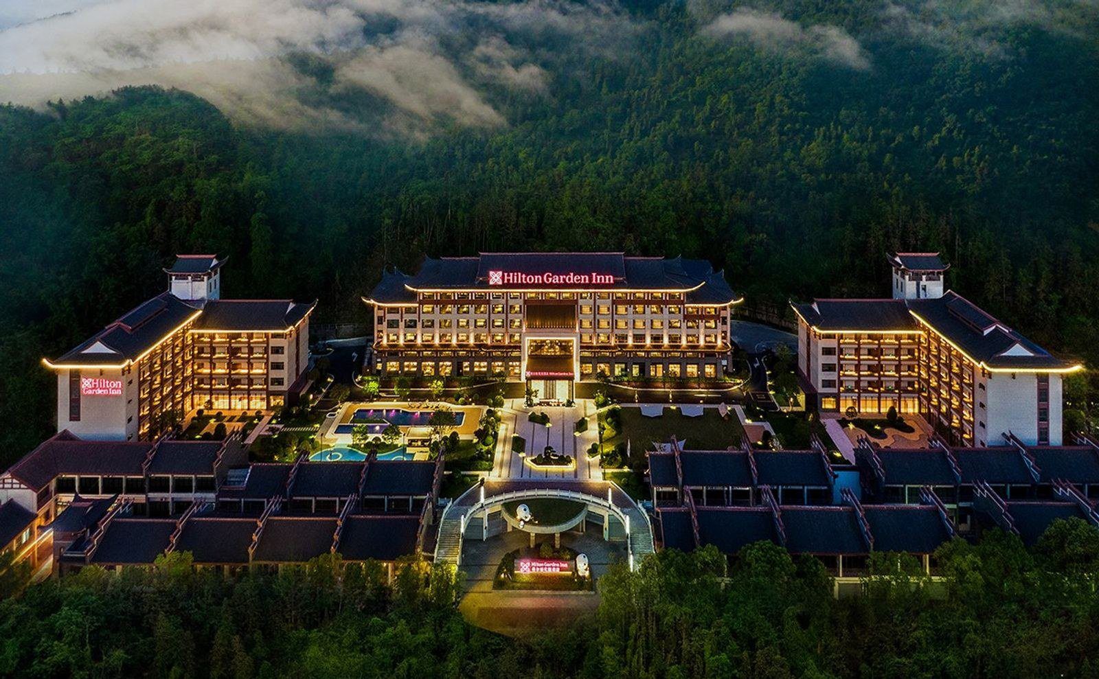 夜間正面空拍的張家界武陵源希爾頓花園酒店,前方是拱形的入口門樓與亮燈步道,後方主樓與兩側樓群環抱庭院和泳池,背景是雲霧下的森林