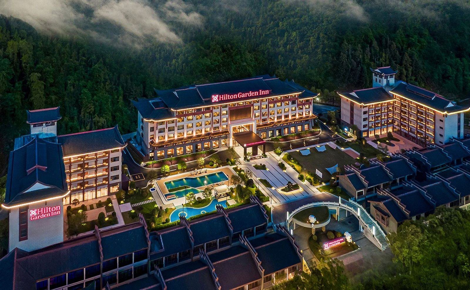 暮色中空拍張家界武陵源希爾頓花園酒店的庭院，主樓與兩側樓群之間是亮燈的戶外泳池、草坪與步道，四周是雲霧下的森林山坡