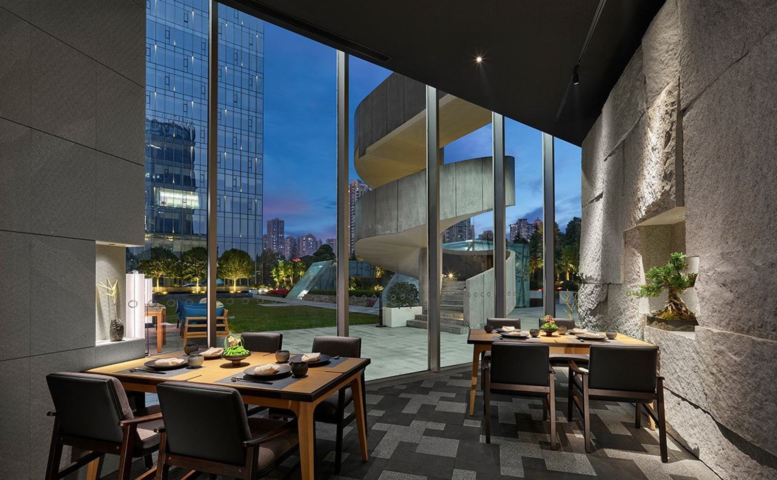 貴陽東景希爾頓酒店燈 AKARI 日料的用餐區，木桌與深色椅子旁是粗石牆，落地窗外是草坪與旋轉樓梯