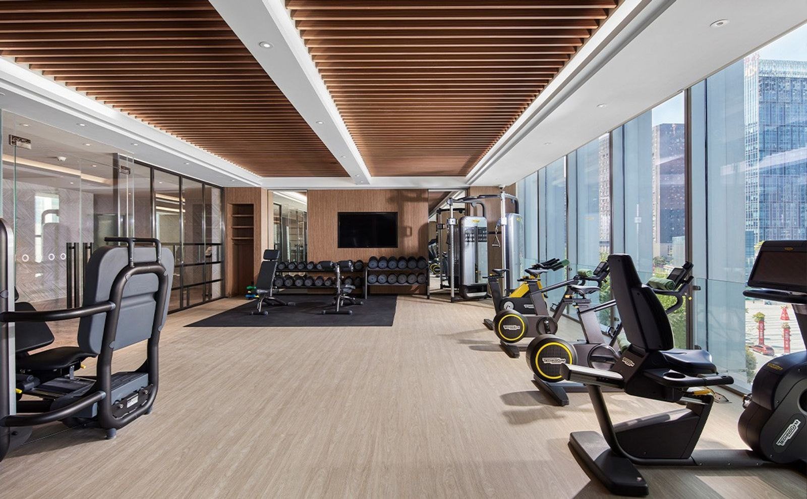貴陽東景希爾頓酒店健身中心，木條天花板下擺著 Technogym 飛輪與重訓器材，右側落地窗外是高樓