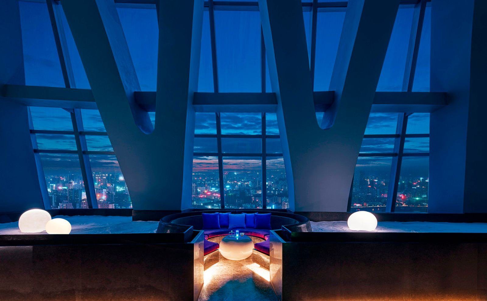 夜晚的空中酒吧，V 形斜柱之間是大片玻璃窗與城市燈火，中央下沉座位區擺著藍色軟墊與發光圓桌