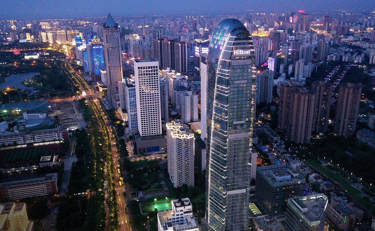 入夜後空拍城市,中央是頂部亮著 Hilton 字樣的弧形高樓,左側是車燈延伸的林蔭大道與周邊樓群