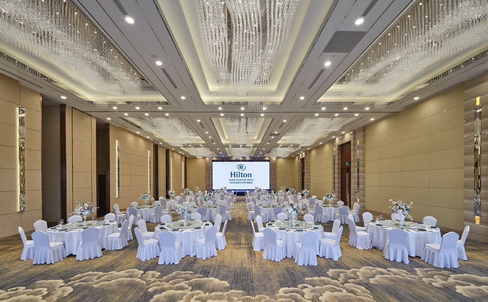 無柱宴會廳排滿圓桌白椅，前方螢幕顯示 Hilton 標誌