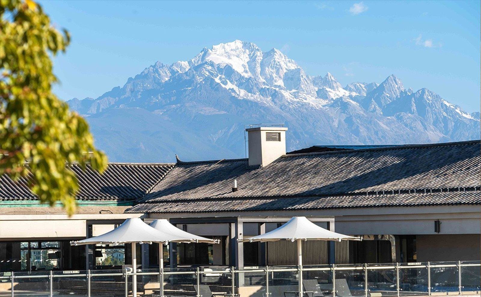 麗江晶璽希爾頓酒店灰瓦屋頂後方的玉龍雪山，前方露台撐著三把白色遮陽傘，四周圍著玻璃欄杆