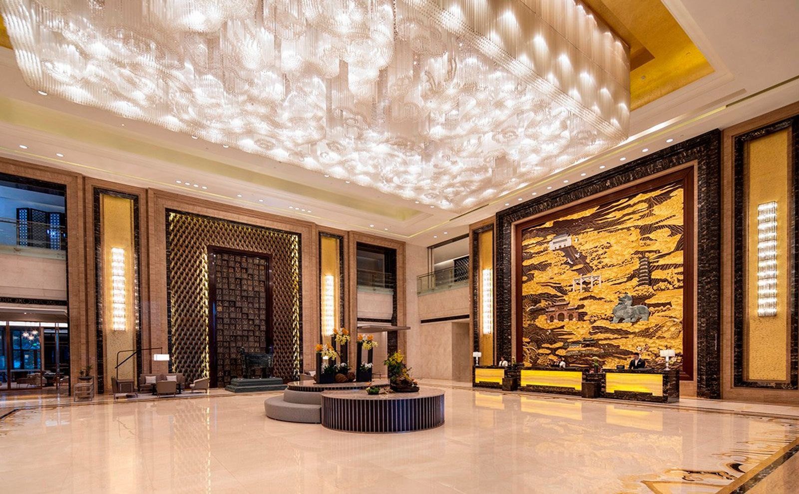 南京希爾頓酒店挑高大堂,天花板垂下大片水晶燈,右側是金色山水浮雕牆前的接待櫃檯,中央是圓形花藝座台