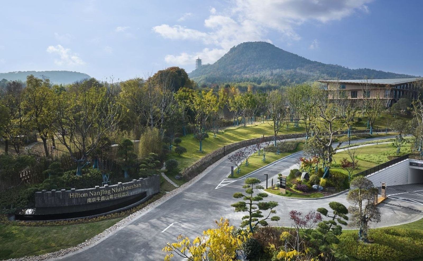 南京牛首山希爾頓酒店入口車道,左側石牆寫著 Hilton Nanjing Niushoushan 館名,後方是綠樹與山頂古塔,右側是酒店建築