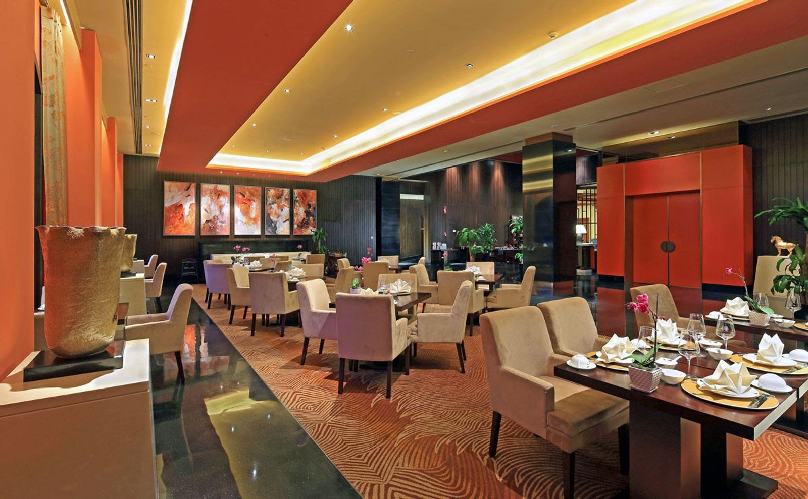青島金沙灘希爾頓酒店膠澳中餐廳大廳，橘紅色牆面掛著抽象畫，擺著米色軟椅與餐桌