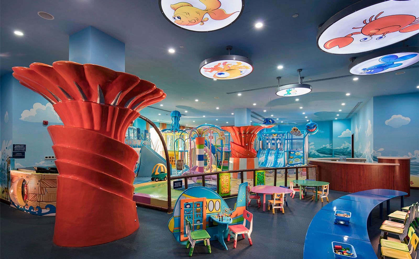 青島金沙灘希爾頓酒店兒童俱樂部，海洋主題壁畫與魚形燈飾下是溜滑梯遊戲區與小桌椅