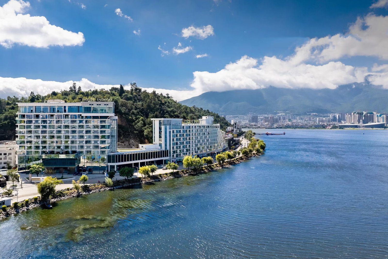 洱海南岸的空拍,左側大樓頂掛著 HOTEL INDIGO 字樣,前方是沿湖綠樹與步道,右後方是下關市區與雲霧中的蒼山
