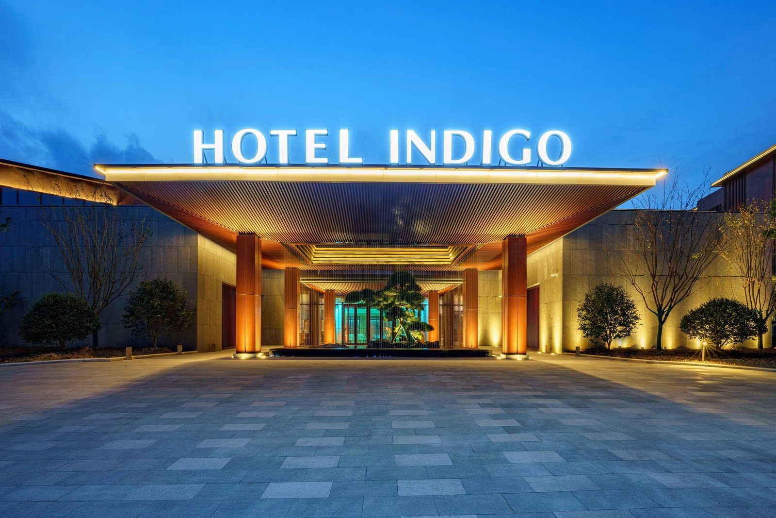 南京園博園英迪格酒店夜間正門,雨遮頂上是 HOTEL INDIGO 招牌,柱廊後方是亮燈的造型松