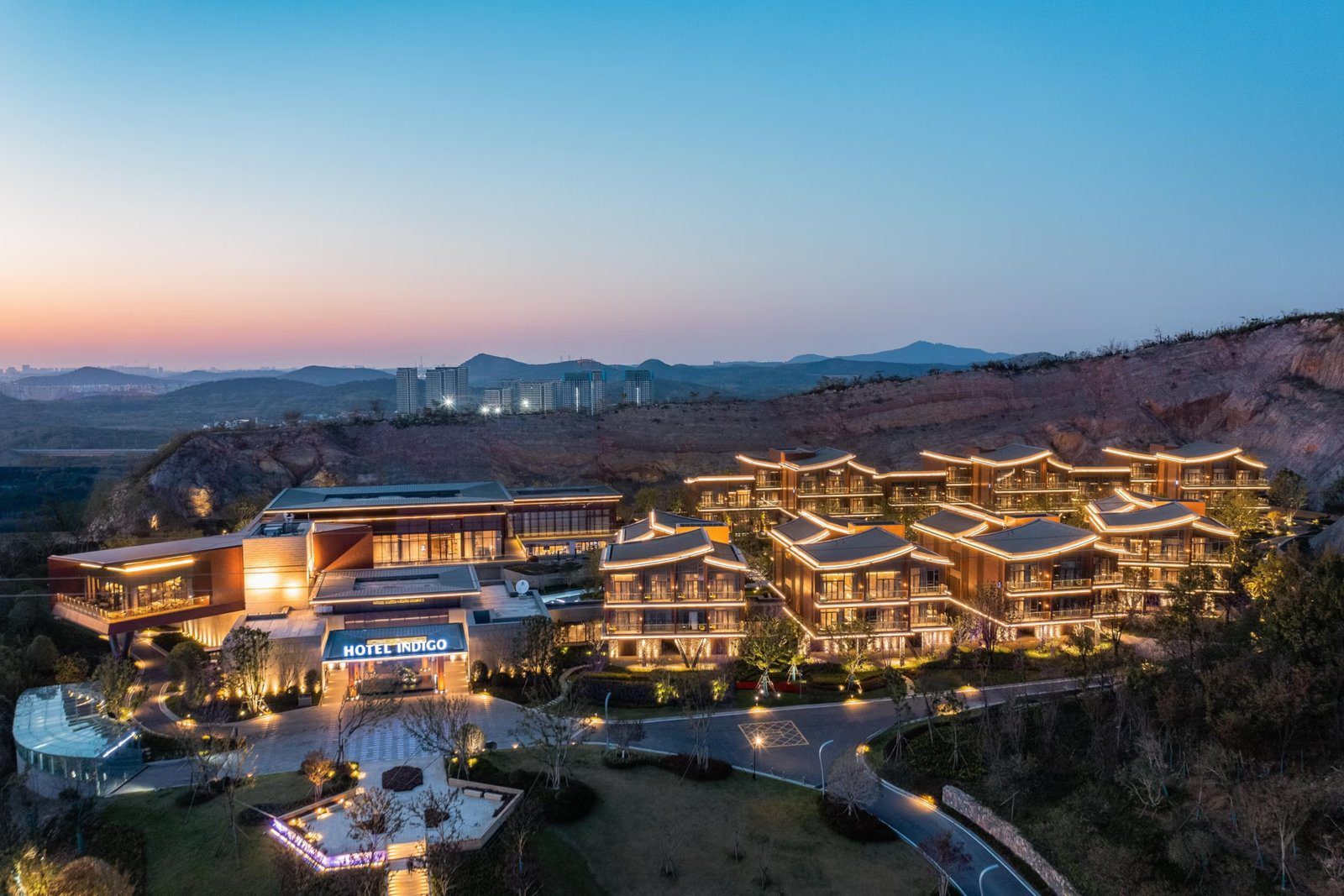 南京園博園英迪格酒店黃昏空拍,入口掛著 HOTEL INDIGO 招牌,右側一排亮燈的別墅沿著礦坑岩壁排開