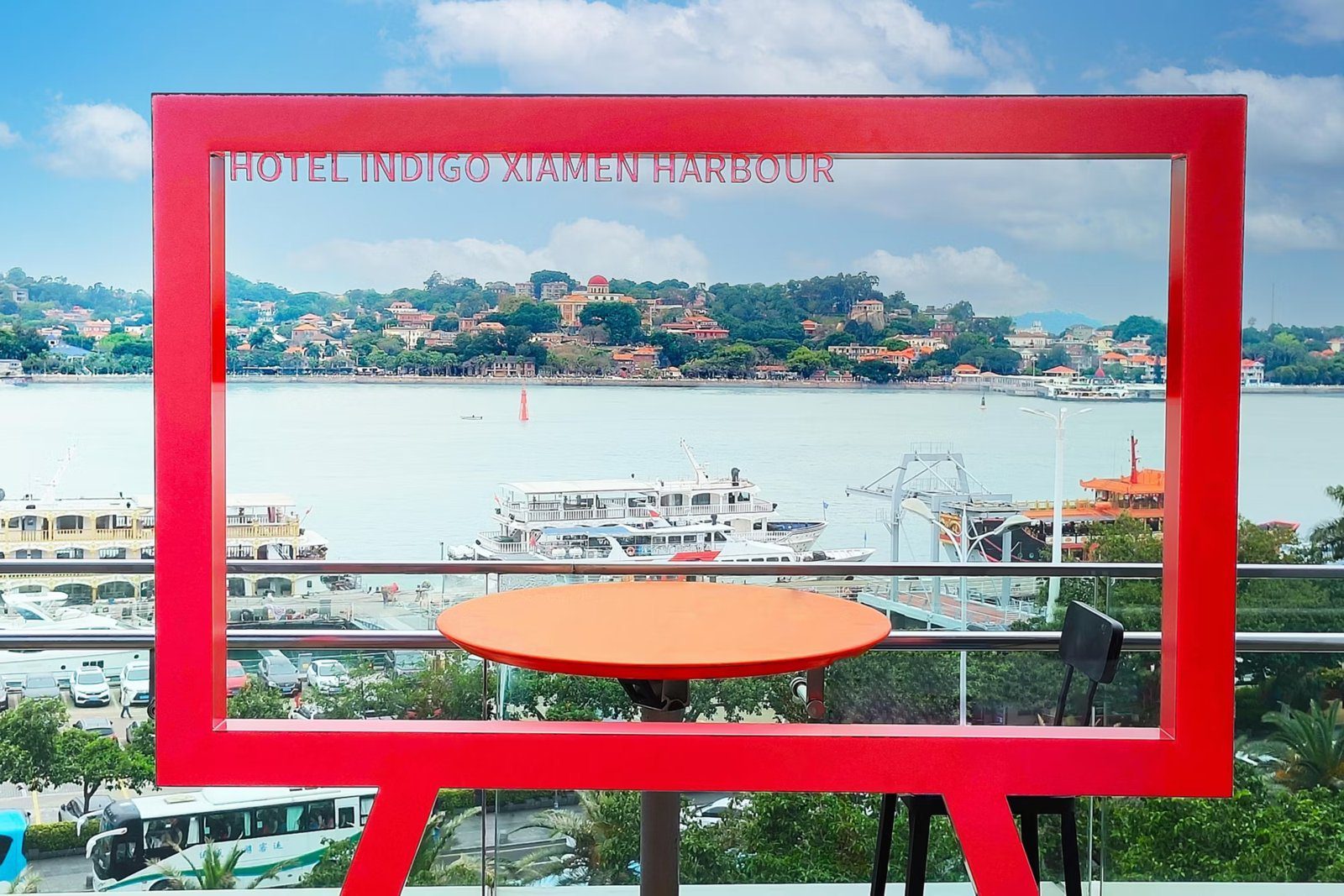 廈門海港英迪格酒店戶外露台上的紅色大相框，框頂刻著 HOTEL INDIGO XIAMEN HARBOUR，框內是海面上停靠的渡輪與對岸紅瓦屋頂的島嶼，前方擺著一張橘色圓桌