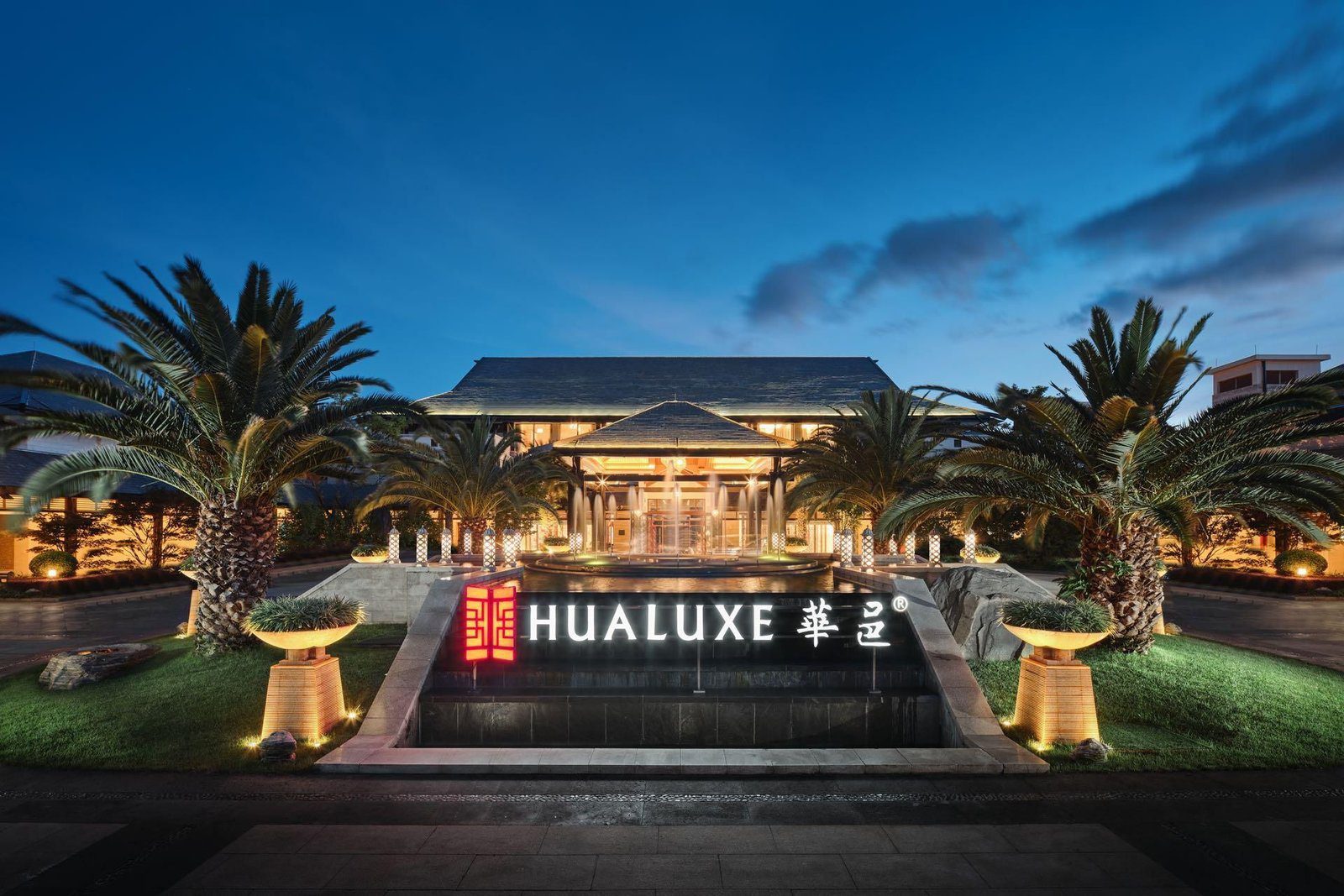 昆明華邑酒店入口夜景,亮燈的 HUALUXE 華邑招牌立在水池前,兩側是棕櫚樹,後方是門廊與噴泉