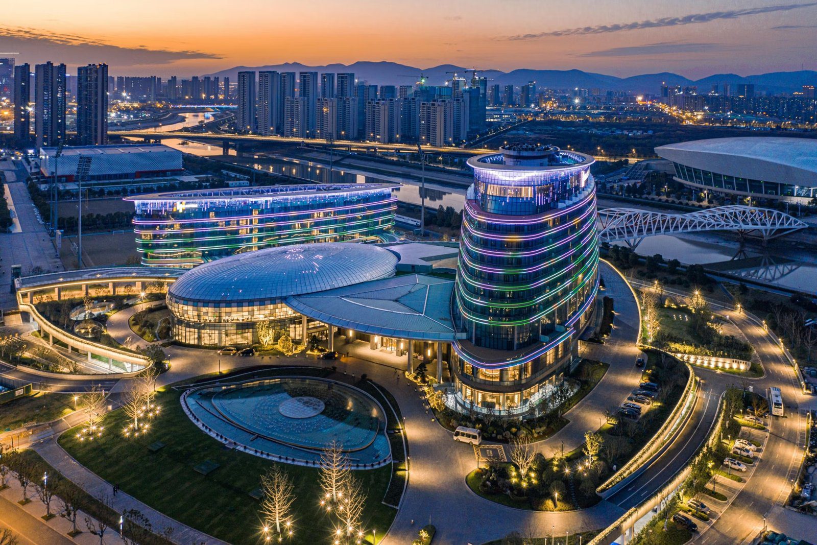 南京長江之舟華邑酒店夜間空拍,圓頂建築與圓柱形塔樓亮著彩色燈帶,右後方是白色桁架橋與體育場館