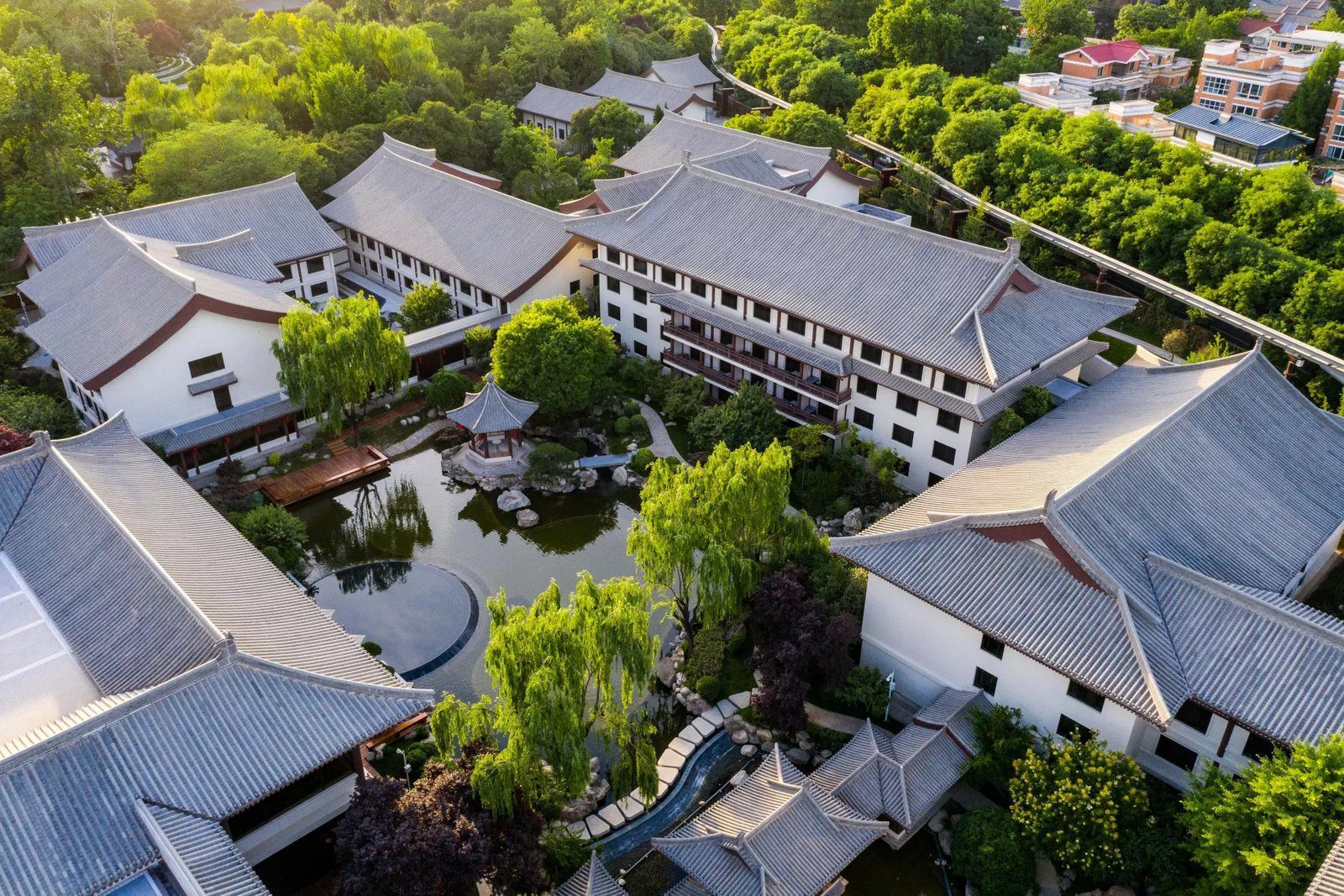 白天空拍西安唐華華邑酒店，灰瓦坡頂的白牆建築圍出中庭，中庭有水池、八角涼亭、木平台與柳樹，四周是茂密的綠樹