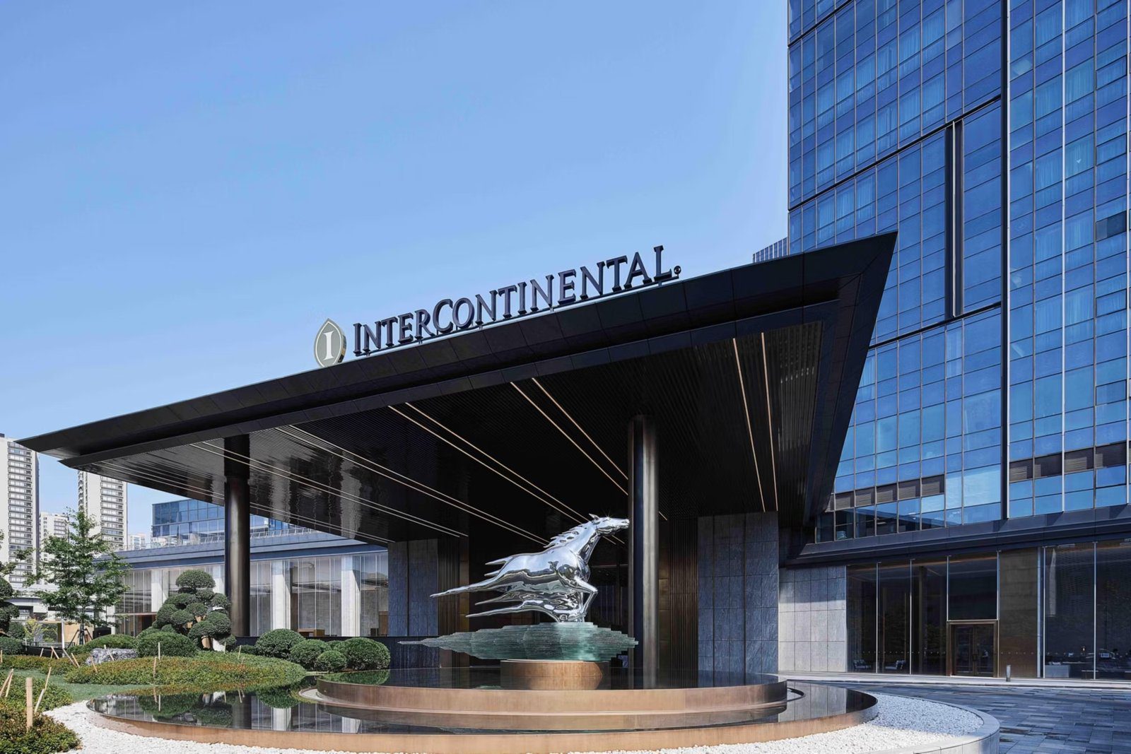 杭州良渚洲際酒店白天的入口雨遮與 INTERCONTINENTAL 招牌,門前是銀色駿馬雕塑與造型松