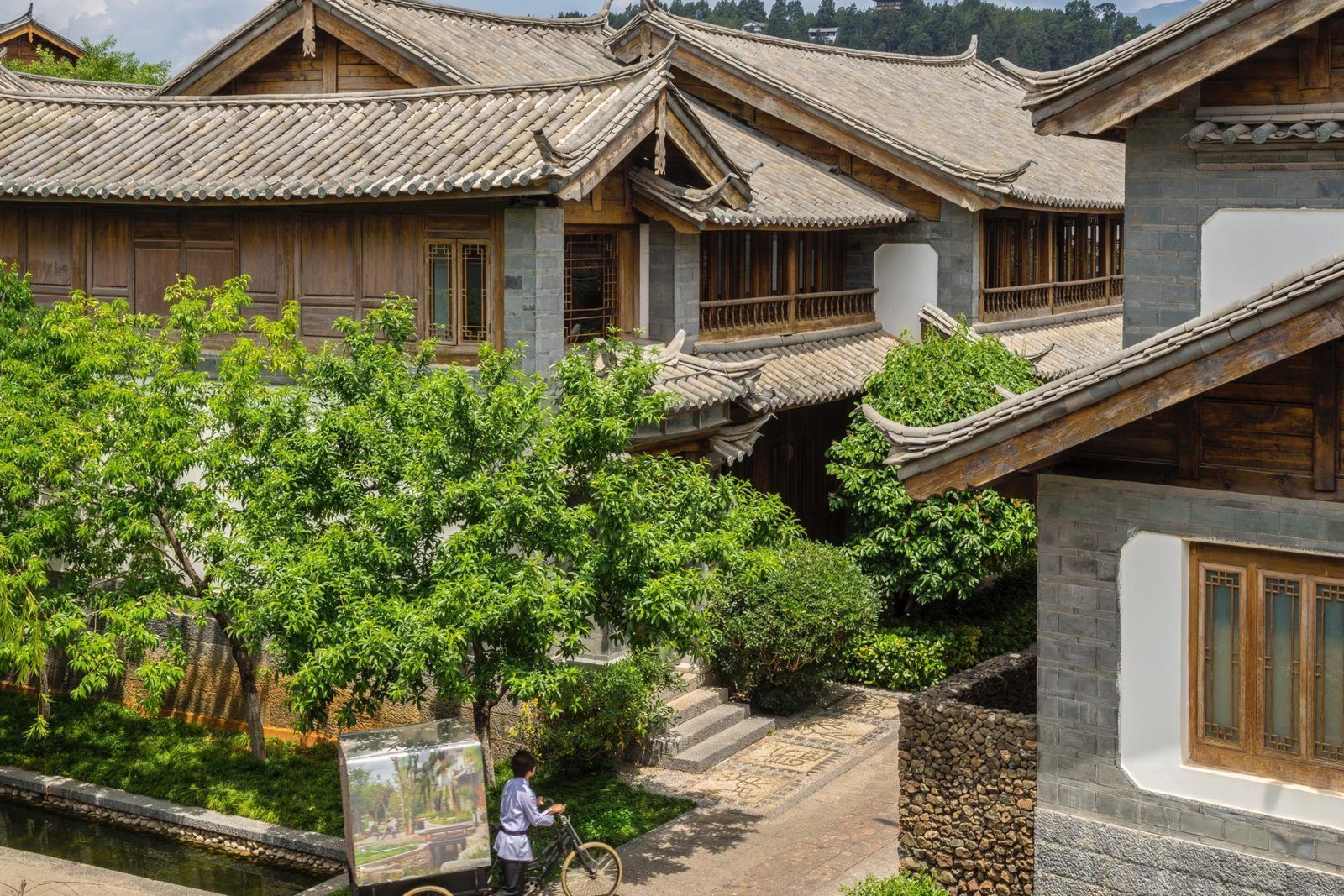 館內的木造二層樓房與灰瓦屋頂，前方綠樹旁是小水渠與石板步道，一名員工推著三輪車經過