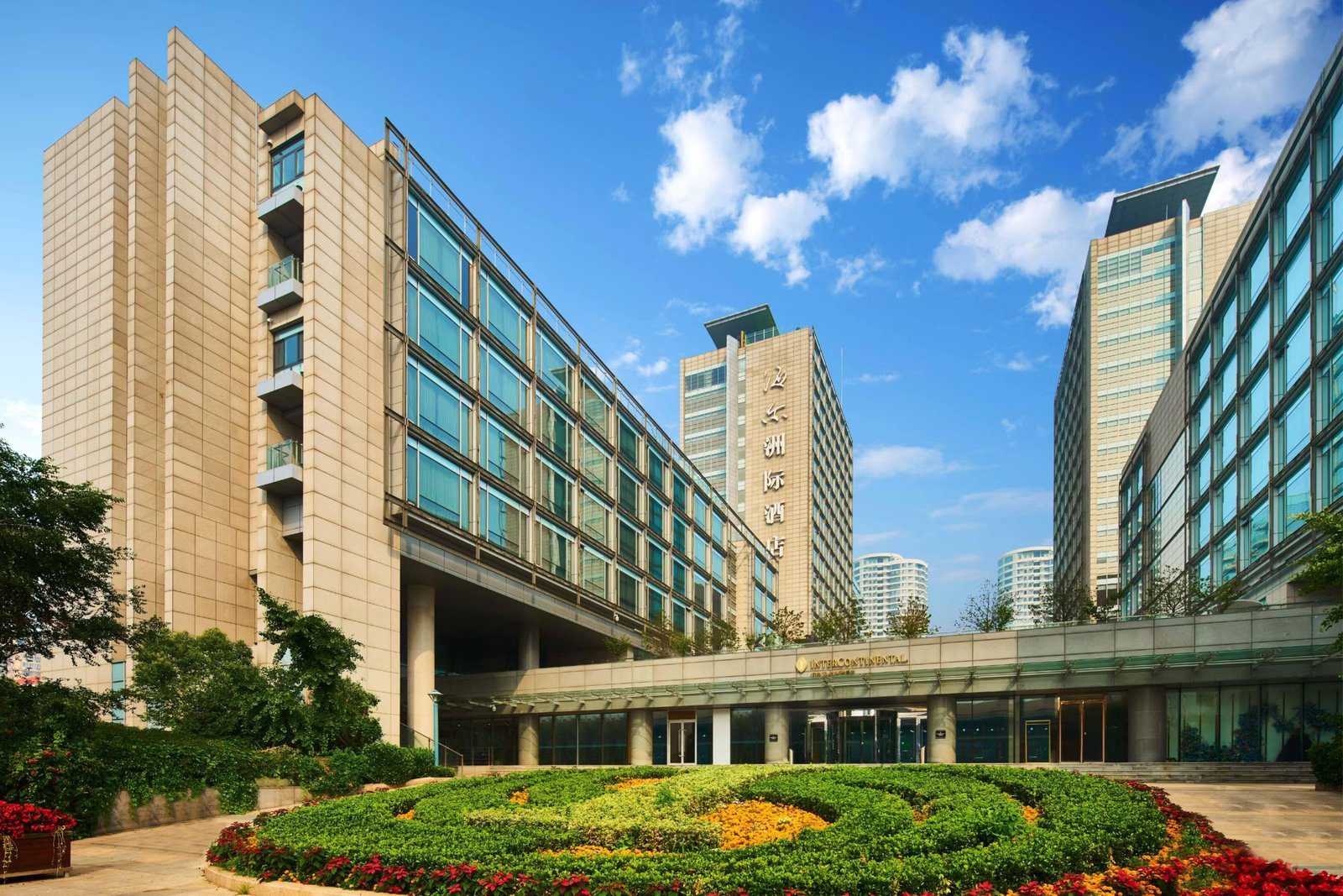 青島海爾洲際酒店白天西入口，雨遮上掛著 InterContinental 字樣，前方是修剪成圖案的花圃