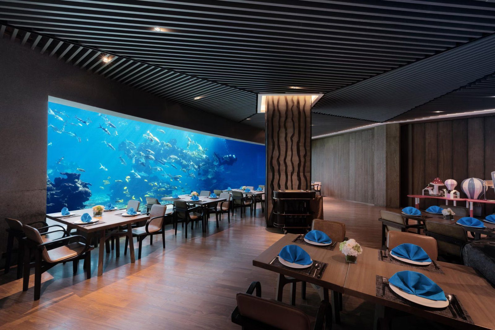 餐廳一整面牆是大型海水缸，魚群與潛水員在珊瑚礁間游動，前方木地板上排著擺好藍色餐巾的餐桌