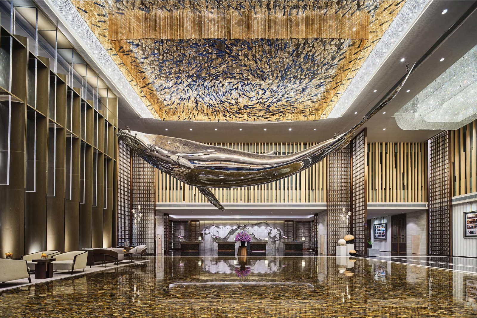 廈門海景洲際酒店挑高大堂，銀色金屬鯨魚雕塑懸吊在半空，上方天花是成群藍色魚形吊飾與金色光帶，下方光亮的石材地面倒映著接待櫃台與紫色花藝